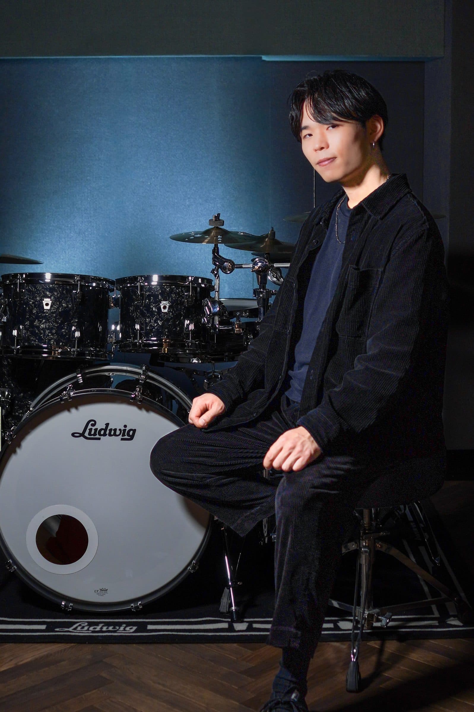 青い照明のスタジオでドラムセットの前に座る木村蓮翔
